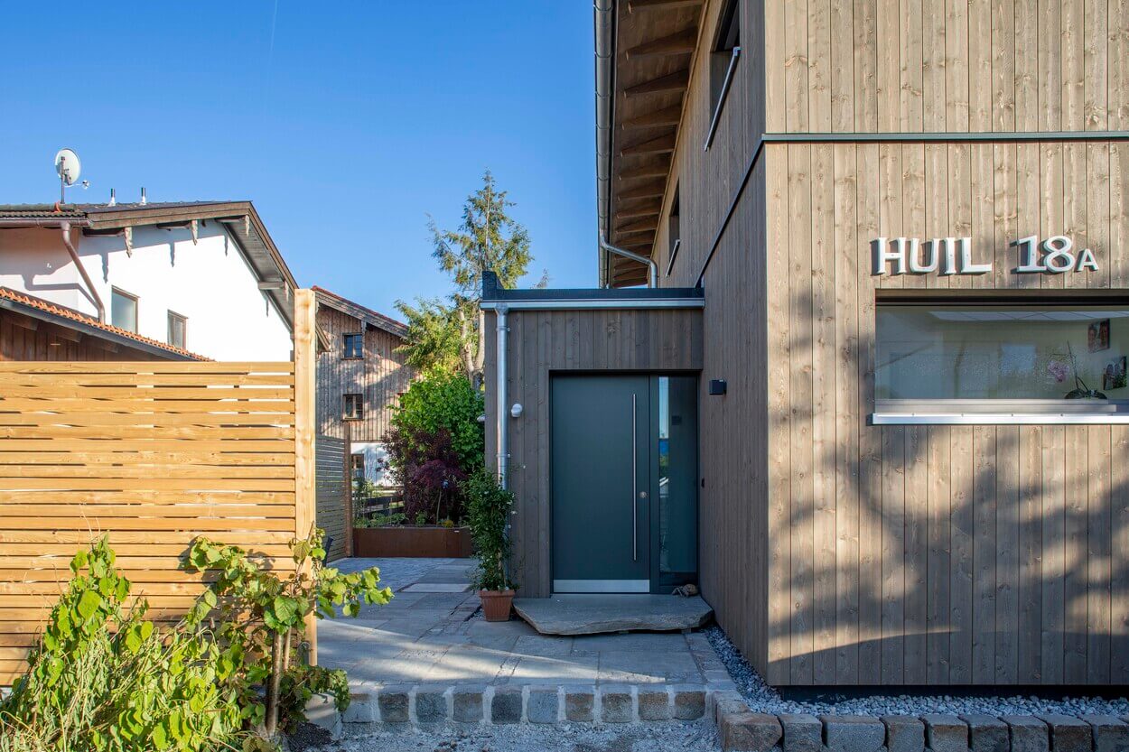 Der Eingang des modernen Holzhauses in Otterfing präsentiert eine stilvolle Tür und gepflegte Pflanzen im Vorgarten.