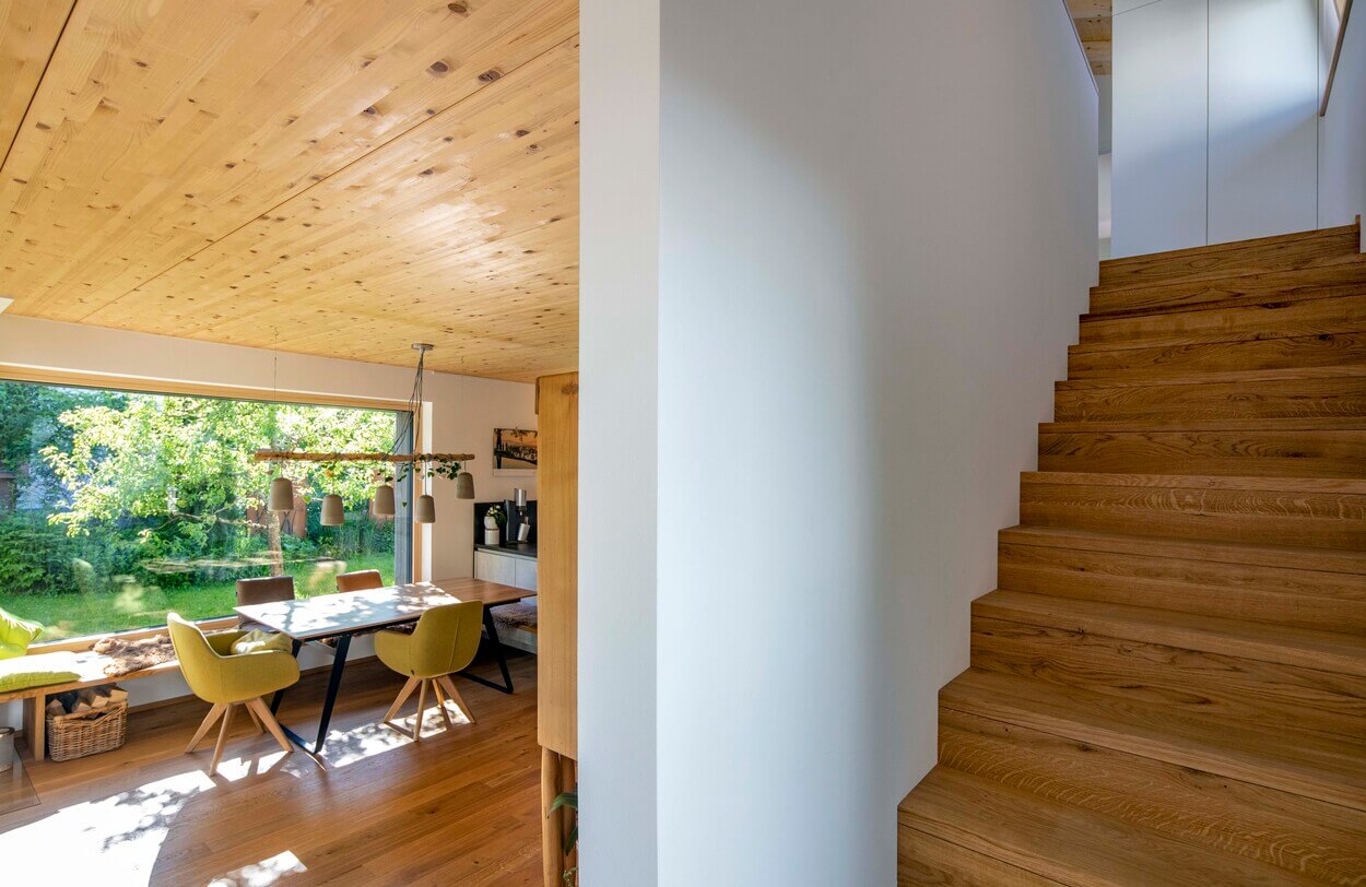 Ein heller Flur im Erdgeschoss eines Holzhauses in Otterfing zeigt eine Treppe und Essbereich mit großen Fenstern.