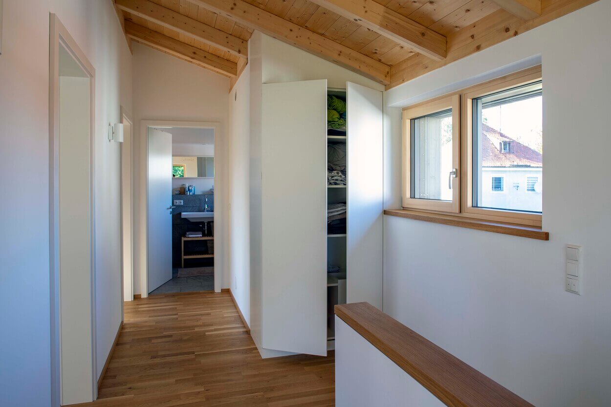 Ein heller Flur in einem Holzhaus in Otterfing zeigt einen offenen Schrank und ein Fenster mit Blick auf ein Nachbarhaus.