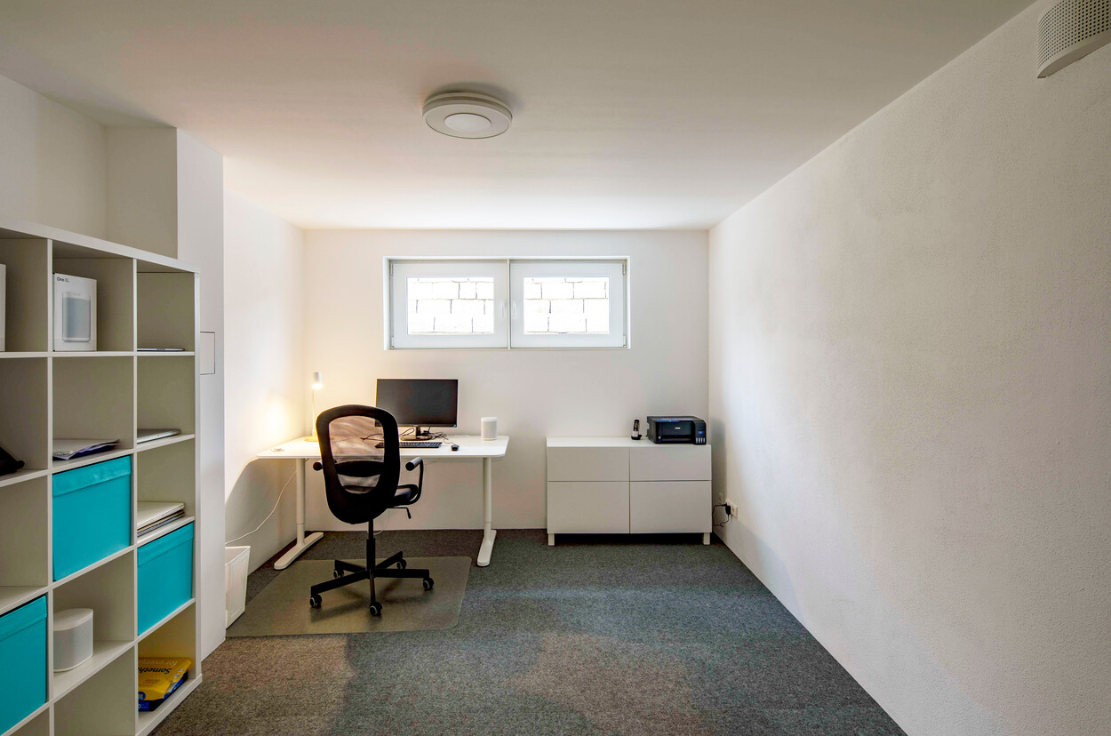 Ein minimalistisches Büro im Passivhaus-Stil mit Schreibtisch und Regalen wird von zwei Fenstern erhellt.