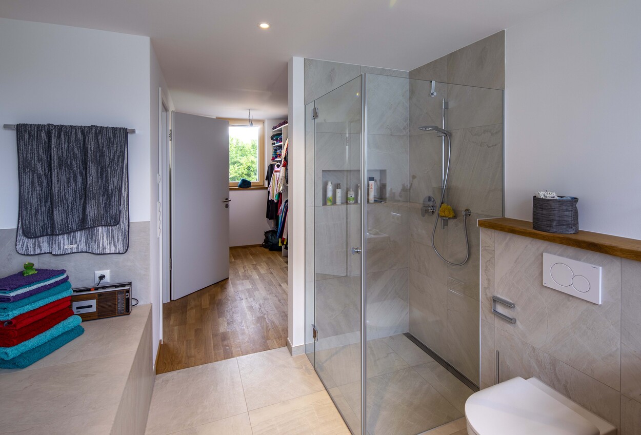 Modernes Badezimmer in einem Holzhaus in Dachau mit Glasdusche und Handtüchern, das einen Blick in den begehbaren Kleiderschrank bietet.