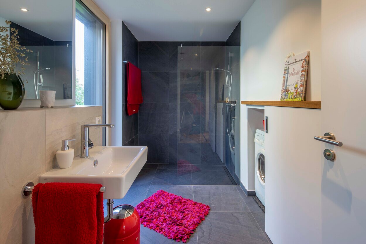 Modernes Badezimmer in einem Holzhaus in Dachau mit Glasdusche, Waschmaschine und roten Akzenten auf Handtuch und Teppich.