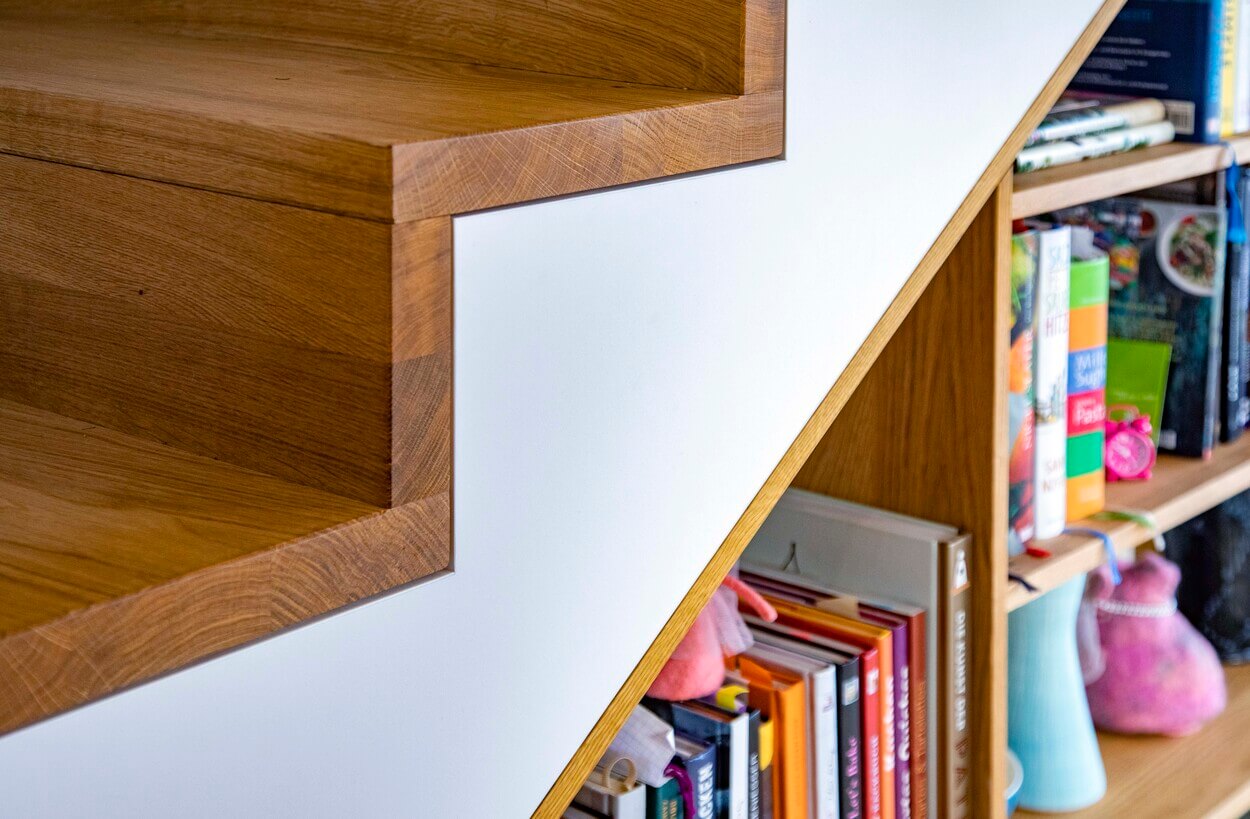 Detail einer Holztreppe in einem Haus in Dachau, mit integrierten Regalen voller bunter Bücher und Dekoration.
