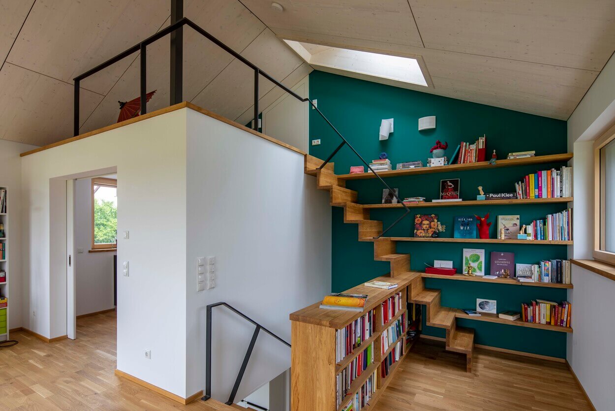 Eine hölzerne Treppe in einem modernen Haus in Dachau führt elegant an einer grünen Wand mit Bücherregalen vorbei.