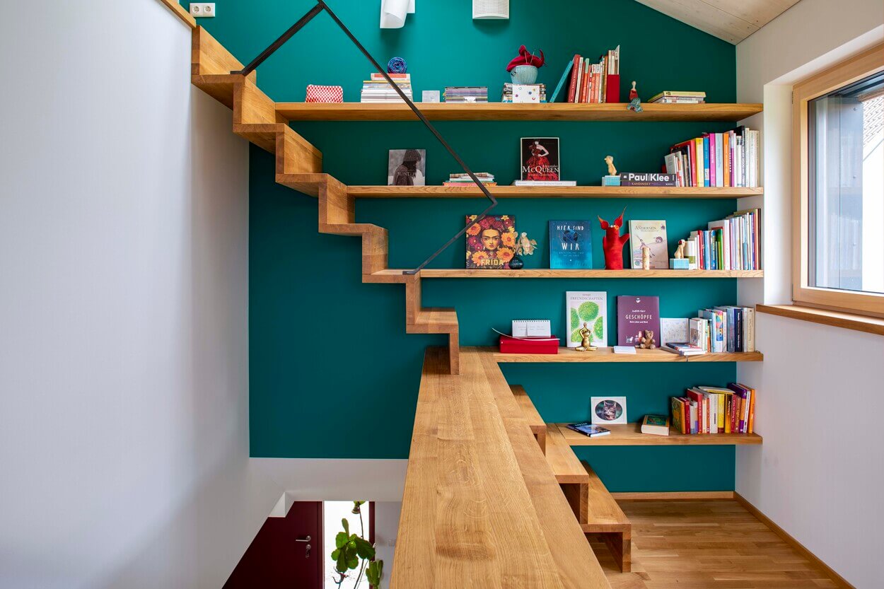 Ein modernes Treppenhaus aus Holz mit integrierten Bücherregalen und grüner Wand verleiht dem Holzhaus in Dachau Charme.