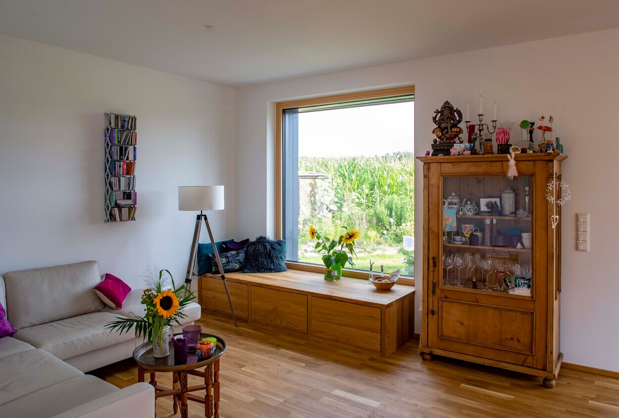 Ein gemütliches Wohnzimmer mit Holzakzenten, großen Fenstern und heller Couch lädt zum Entspannen im Holzhaus Dachau ein.