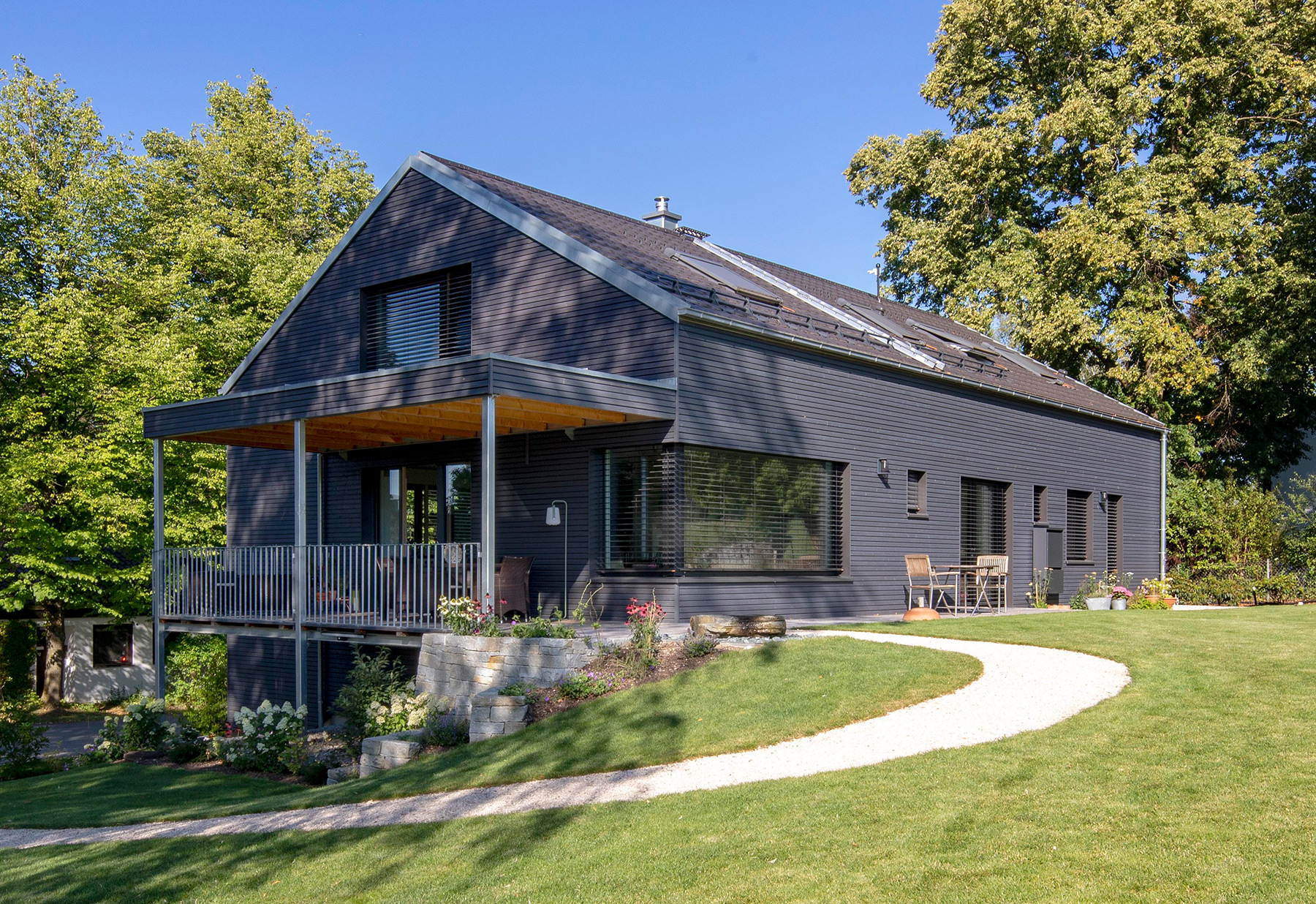 Ein modernes Holzhaus in Glonn steht von Bäumen umgeben auf einem gepflegten Rasen mit geschwungenem Weg.