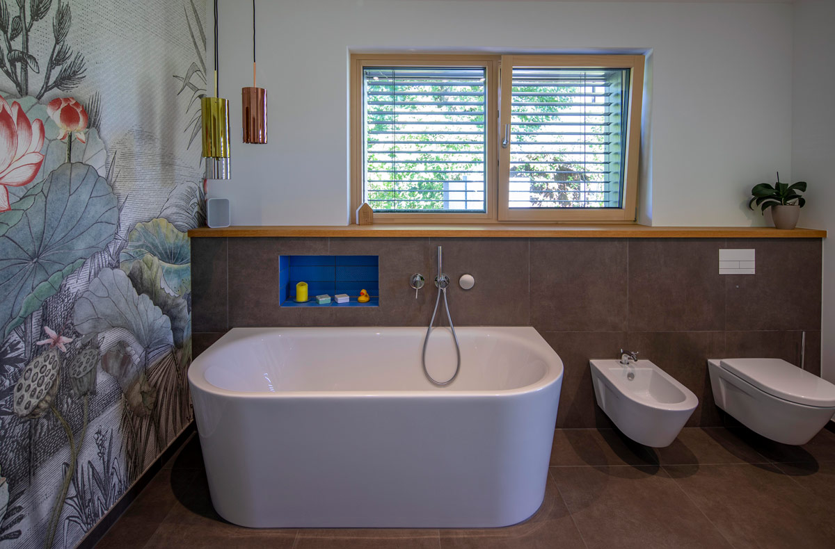 Ein modernes Badezimmer in einem Holzhaus in Glonn zeigt eine freistehende Badewanne vor einem floralen Wandbild und großem Fenster.