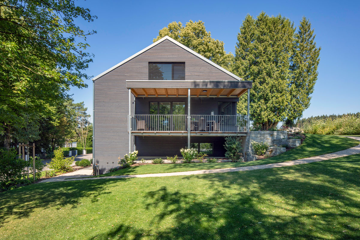 Ein modernes Holzhaus in Glonn mit Balkon steht umgeben von üppigem Grün und Bäumen unter strahlend blauem Himmel.