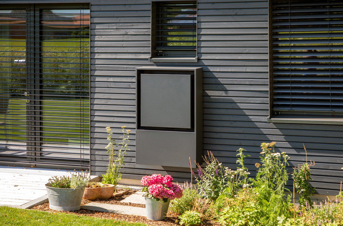 Ein modernes Holzhaus in Glonn mit grauer Fassade und großen Fenstern wird von blühenden Pflanzen im Vordergrund umrahmt.