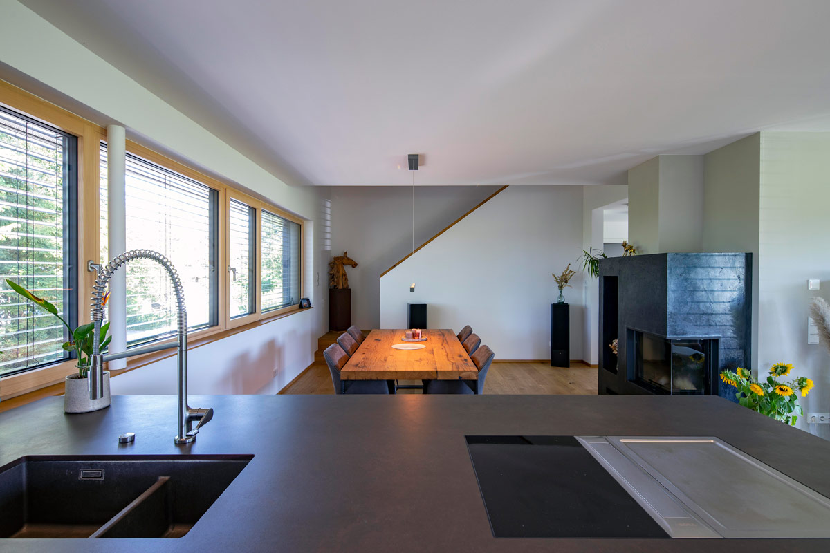 Im modernen Esszimmer des Holzhauses in Glonn steht ein großer Holztisch mit Stühlen, umgeben von Fensterfronten und einem Kamin.