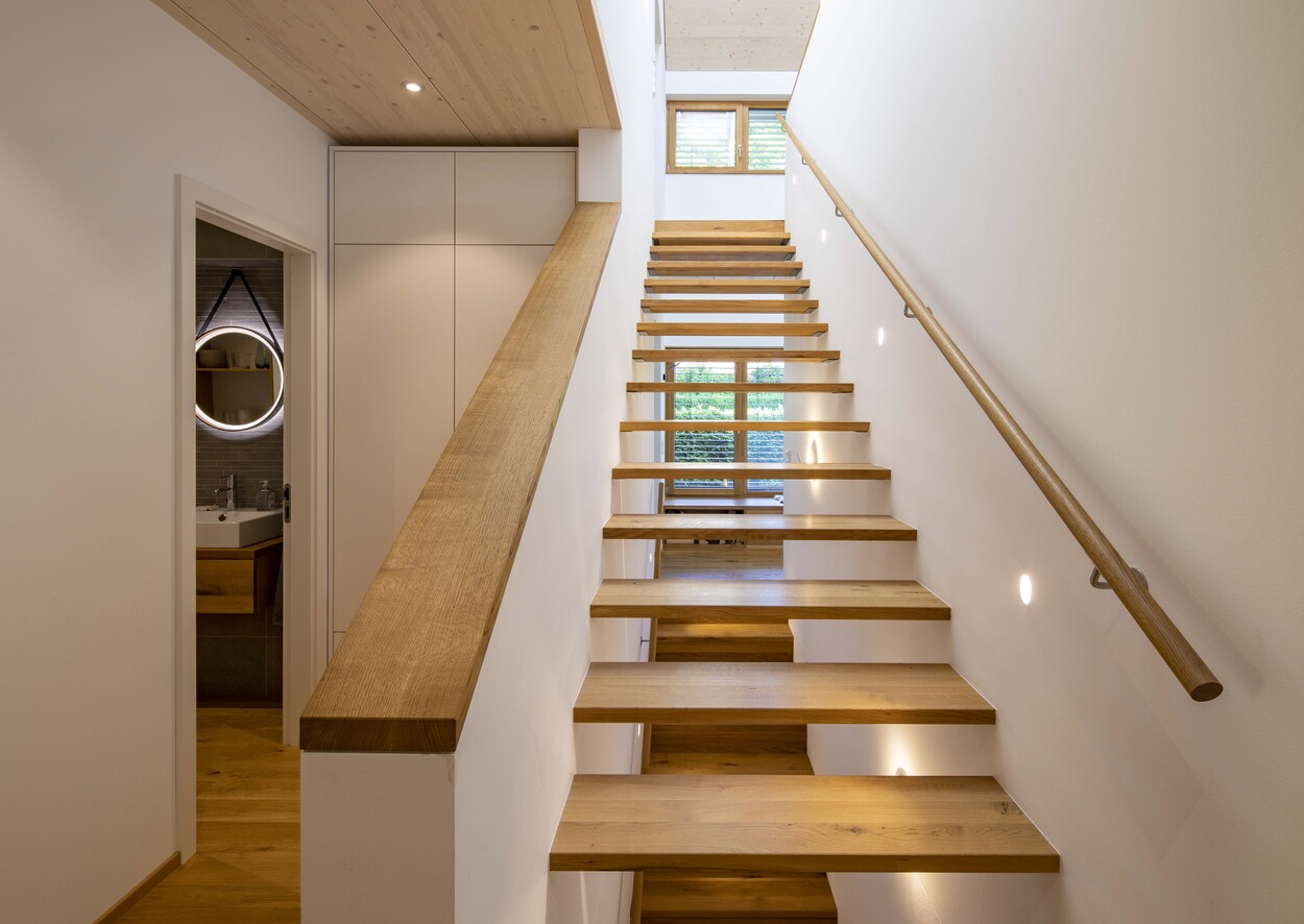 Eine moderne Holztreppe führt in einem Groebenzell-Haus zu einem hellen, minimalistischen Obergeschoss mit Fenstern.