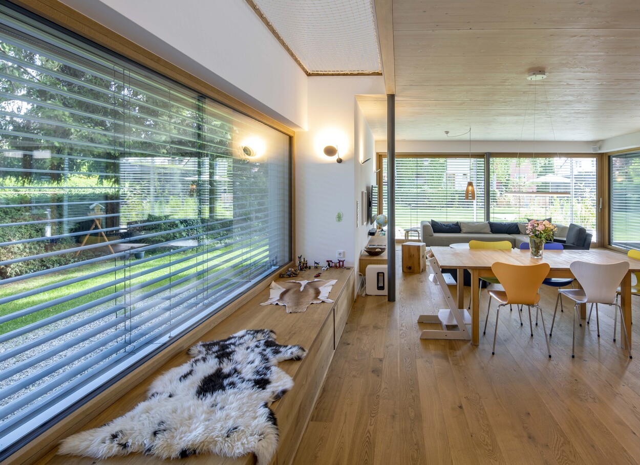 Ein modernes Holzhaus in Gröbenzell präsentiert ein gemütliches Wohnzimmer mit großen Fenstern und einem eleganten Essbereich.