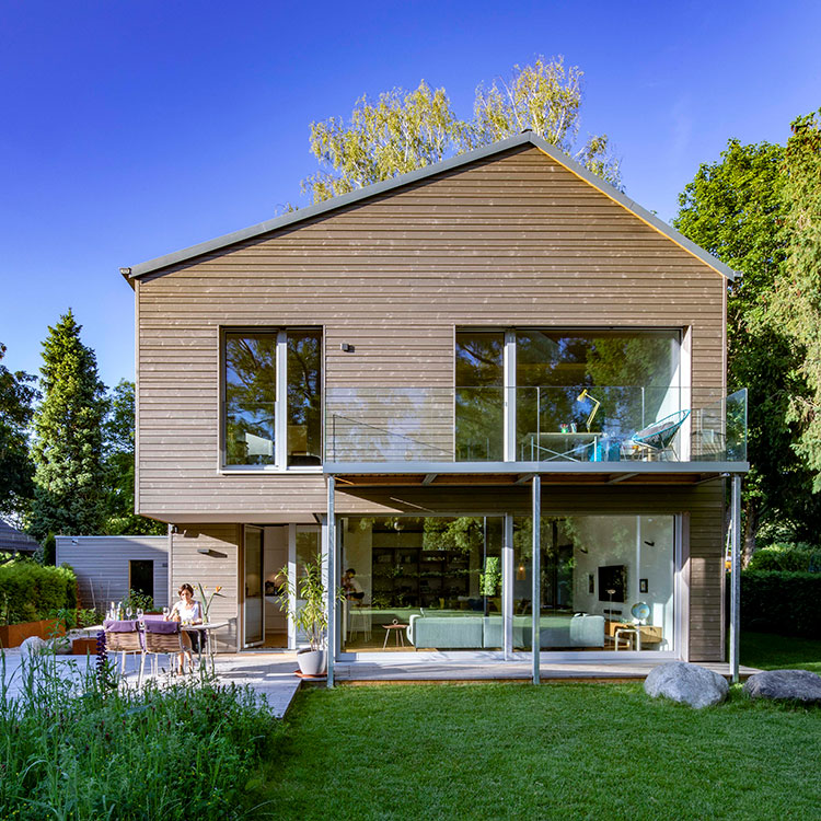 Ein modernes Holzhaus in Gröbenzell mit großen Glasfronten steht in einem grünen Garten unter strahlend blauem Himmel.