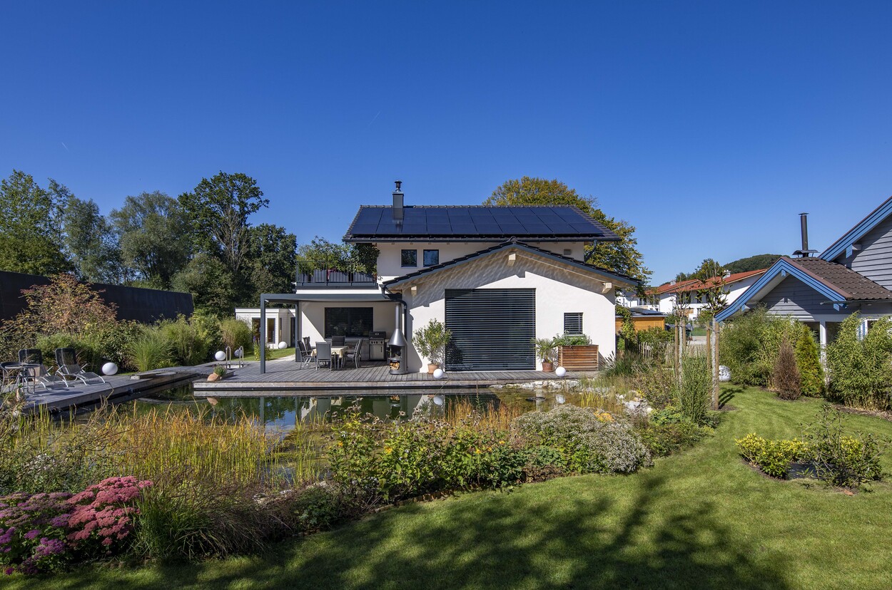 Ein modernes Einfamilienhaus in Prien mit Solardach steht in einem gepflegten Garten neben einem idyllischen Naturteich.