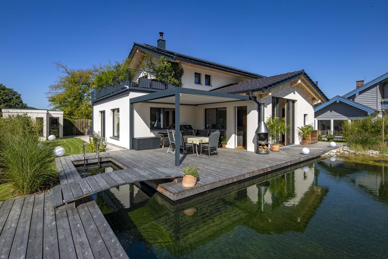 Ein modernes Einfamilienhaus in Prien mit Holzterrasse und Teich, umgeben von üppigem Grün bei strahlend blauem Himmel.