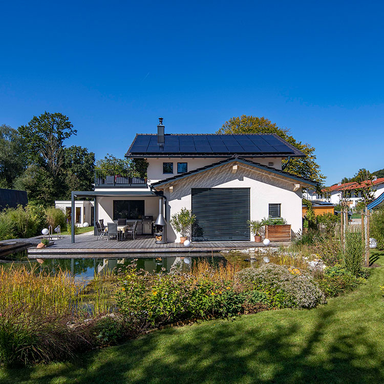 Ein modernes Einfamilienhaus in Prien mit Solarpanelen und üppigem Garten erstrahlt unter klarem blauem Himmel.