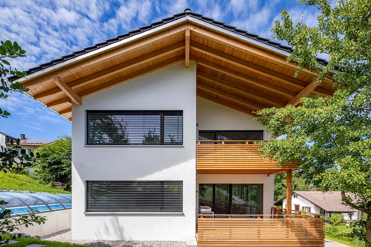 Ein modernes Holzhaus in Rimsting mit großen Fenstern und Balkon, umgeben von grünen Bäumen unter blauem Himmel.