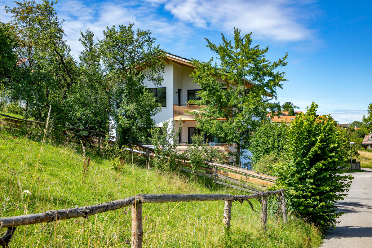 Ein modernes Holzhaus in Rimsting thront auf einem sanften Hügel, umgeben von üppigem Grün und einem strahlenden Himmel.