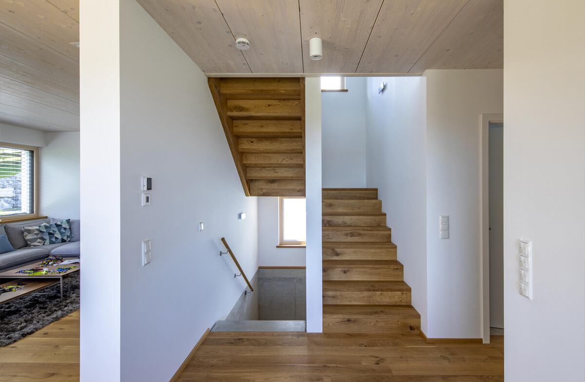 Ein modernes Holzhaus in Rimsting präsentiert ein helles Treppenhaus mit eleganter Holztreppe und angrenzendem Wohnzimmer.