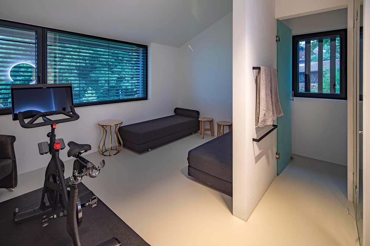 In einem modernen Raum stehen ein Heimtrainer und eine schwarze Liege neben großen Fenstern mit Jalousien.