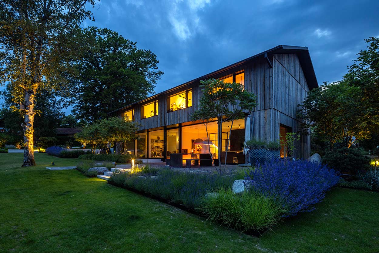 Ein modernes Holzhaus strahlt warmes Licht aus, umgeben von einem gepflegten Garten bei Dämmerung.