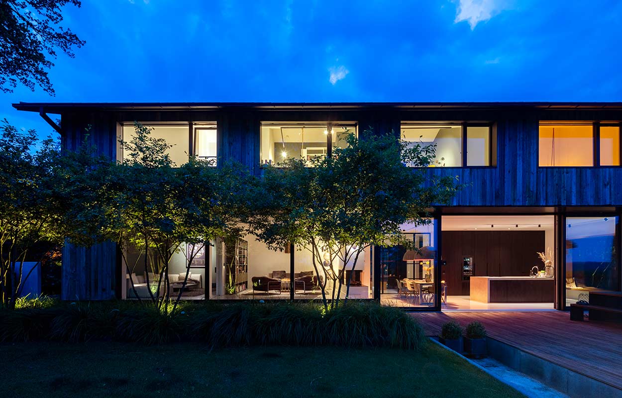 Ein modernes, zweistöckiges Haus mit großen Fenstern strahlt abends warmes Licht in der Dämmerung aus.