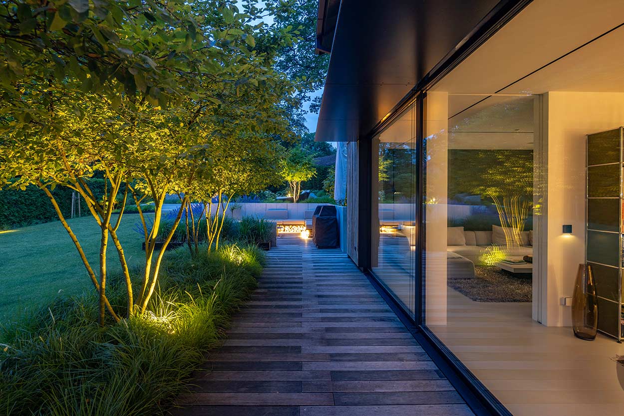 Ein beleuchteter Gartenweg führt an einem modernen Haus mit Glasfassade entlang, umgeben von gepflegtem Rasen und Bäumen.