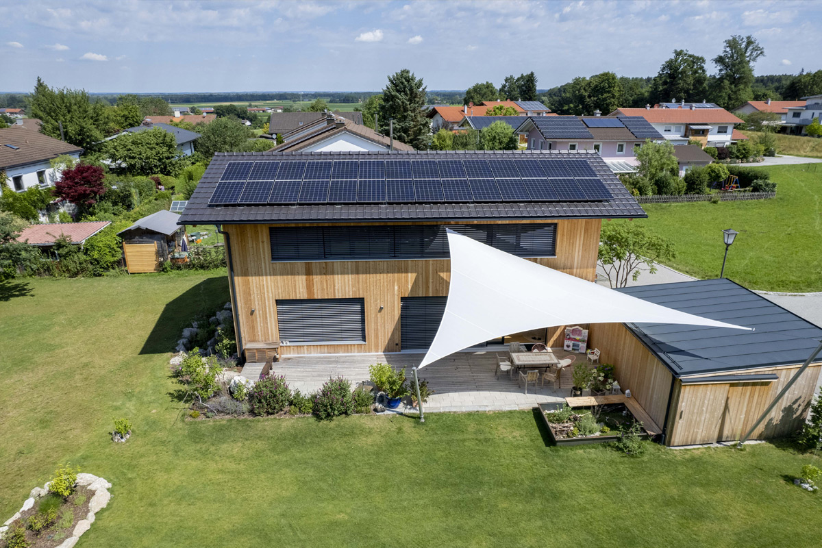 Ein modernes Holzhaus in Neubeuern mit Solarmodulen und einer Terrasse, die von einem eleganten weißen Sonnensegel beschattet wird.