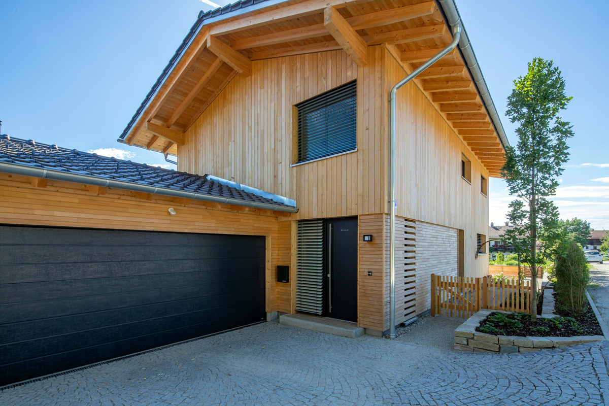Moderne Außenansicht eines Holzhauses in Chieming mit großem Garagentor und gepflegtem Vorgarten an einem sonnigen Tag.