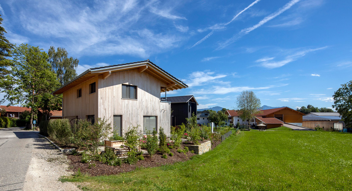 Ein modernes Holzhaus in Chieming steht unter blauem Himmel, umgeben von üppigem Grün und gepflegten Gärten.