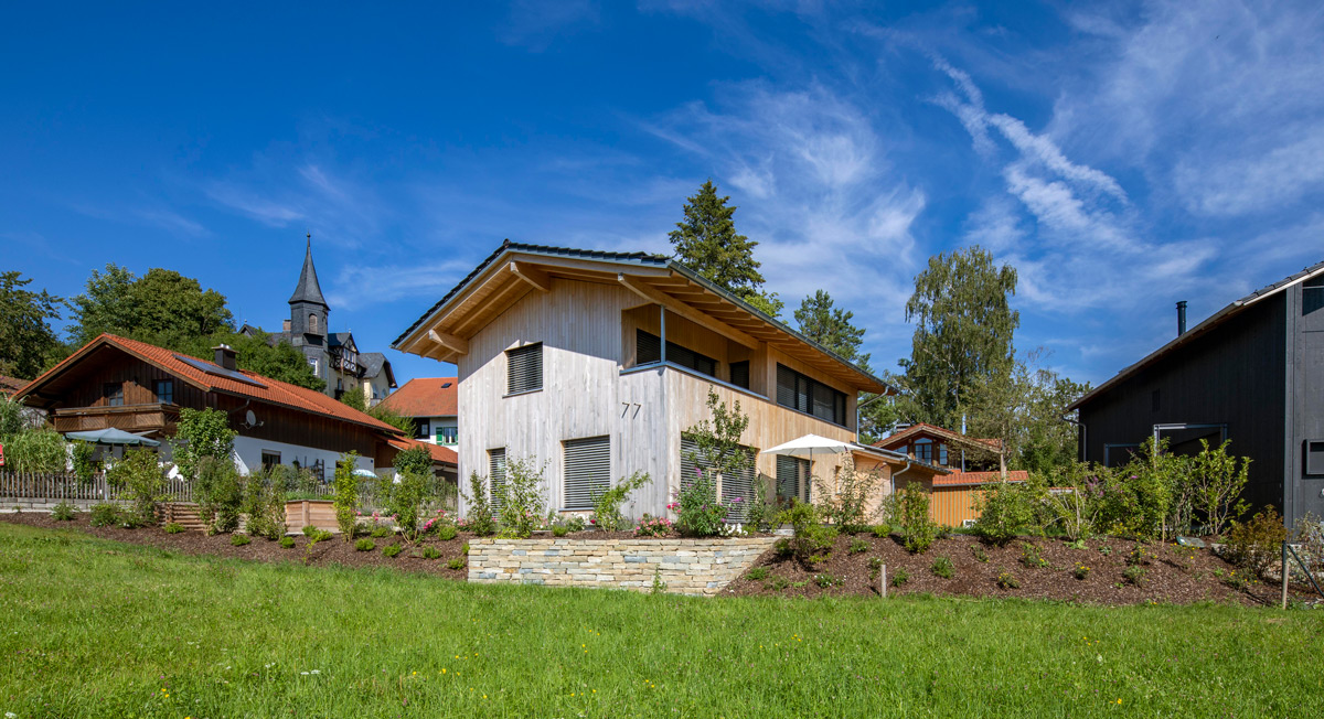 Ein modernes Holzhaus in Chieming steht unter klarem Himmel, umgeben von gepflegten Gärten und traditionellen Gebäuden.