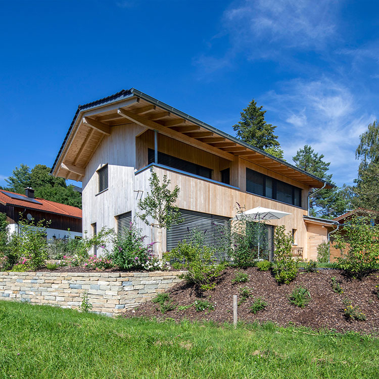 Ein modernes Holzhaus in Chieming steht in einem gepflegten Garten unter strahlend blauem Himmel.