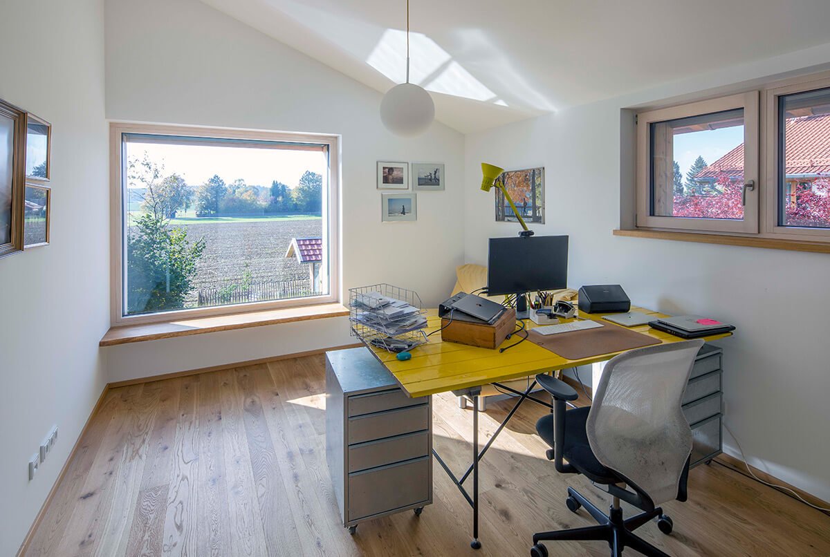 Ein helles Arbeitszimmer in einem Holzhaus in Oberbiberg bietet durch ein großes Fenster Blick auf weite Felder.