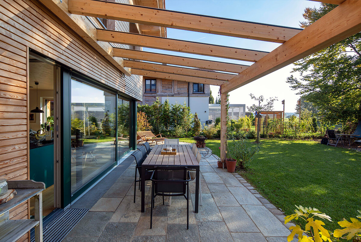 Die Außenansicht eines modernen Holzhauses in Oberbiberg zeigt eine Terrasse mit Pergola und gepflegtem Garten.