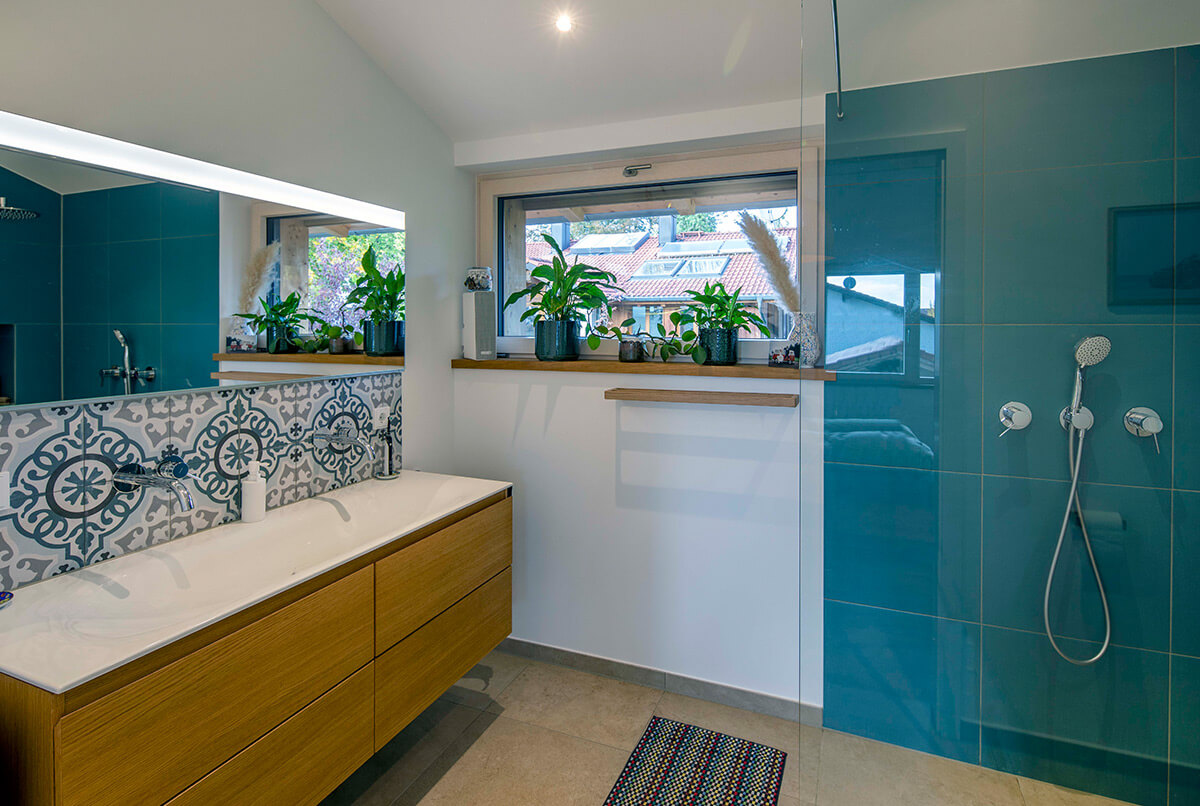 Ein modernes Badezimmer in einem Holzhaus in Oberbiberg mit stilvollen blauen Fliesen und grünen Pflanzen auf dem Fensterbrett.