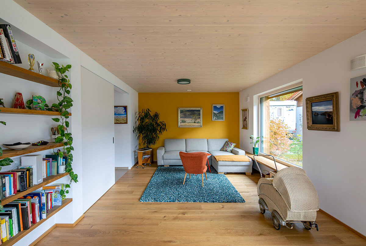 Ein gemütliches Wohnzimmer in Oberbiberg mit Holzdecke, gelber Akzentwand, Sofa und großen Fenstern mit Ausblick.