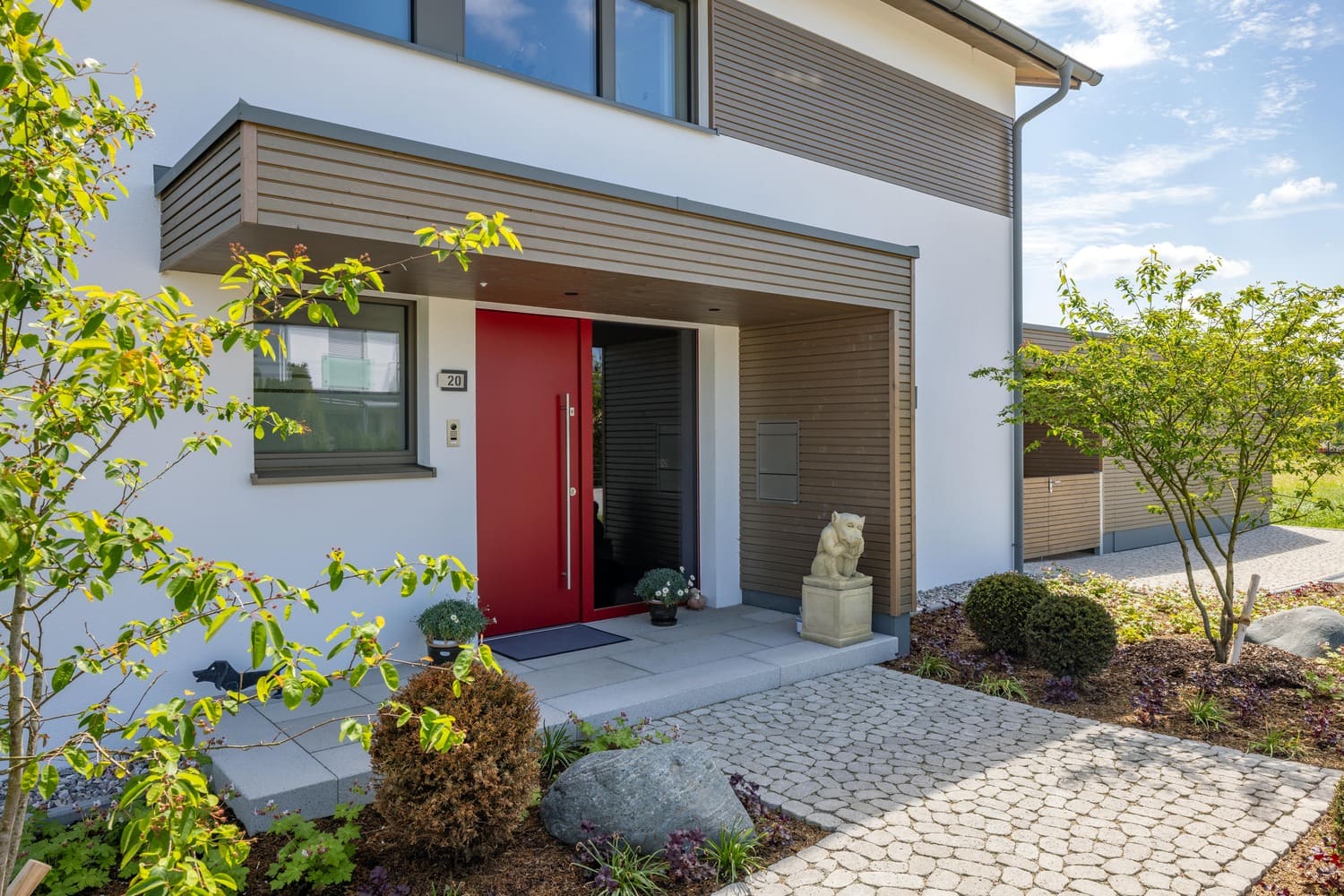 Ein verputztes Holzhaus in Weilheim zeigt eine rote Haustür mit Glaselementen und eine hölzerne Eingangsüberdachung.