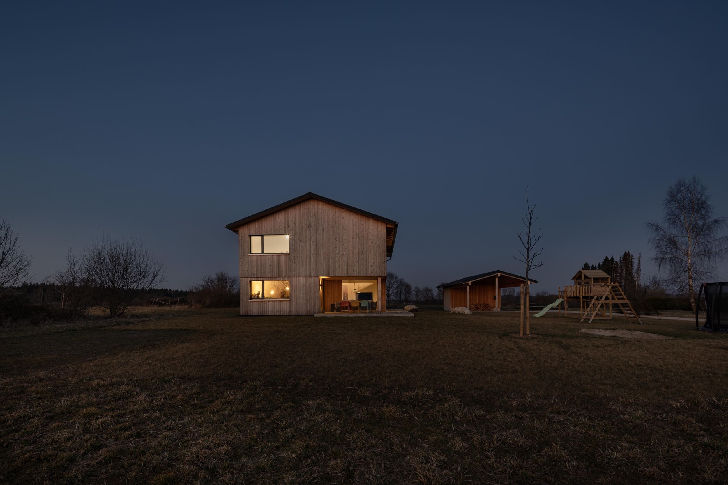 Ein modernes Holzhaus strahlt abends warmes Licht aus, während der umliegende Garten in der ruhigen Dämmerung liegt.