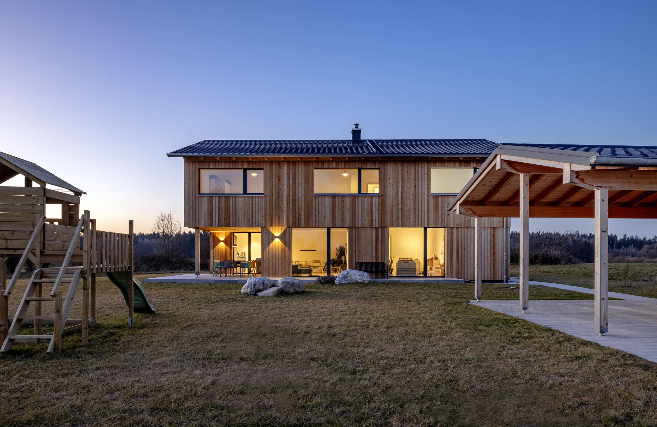 Ein modernes Holzhaus mit beleuchteten Fenstern steht bei Dämmerung neben einem Carport auf einer großen Wiese.