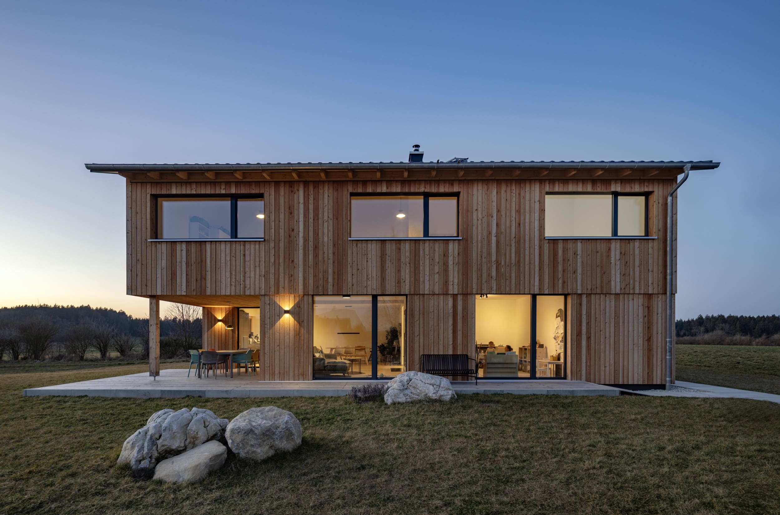 Ein modernes Holzhaus mit großen Fenstern steht in einer ruhigen Landschaft bei Sonnenuntergang.