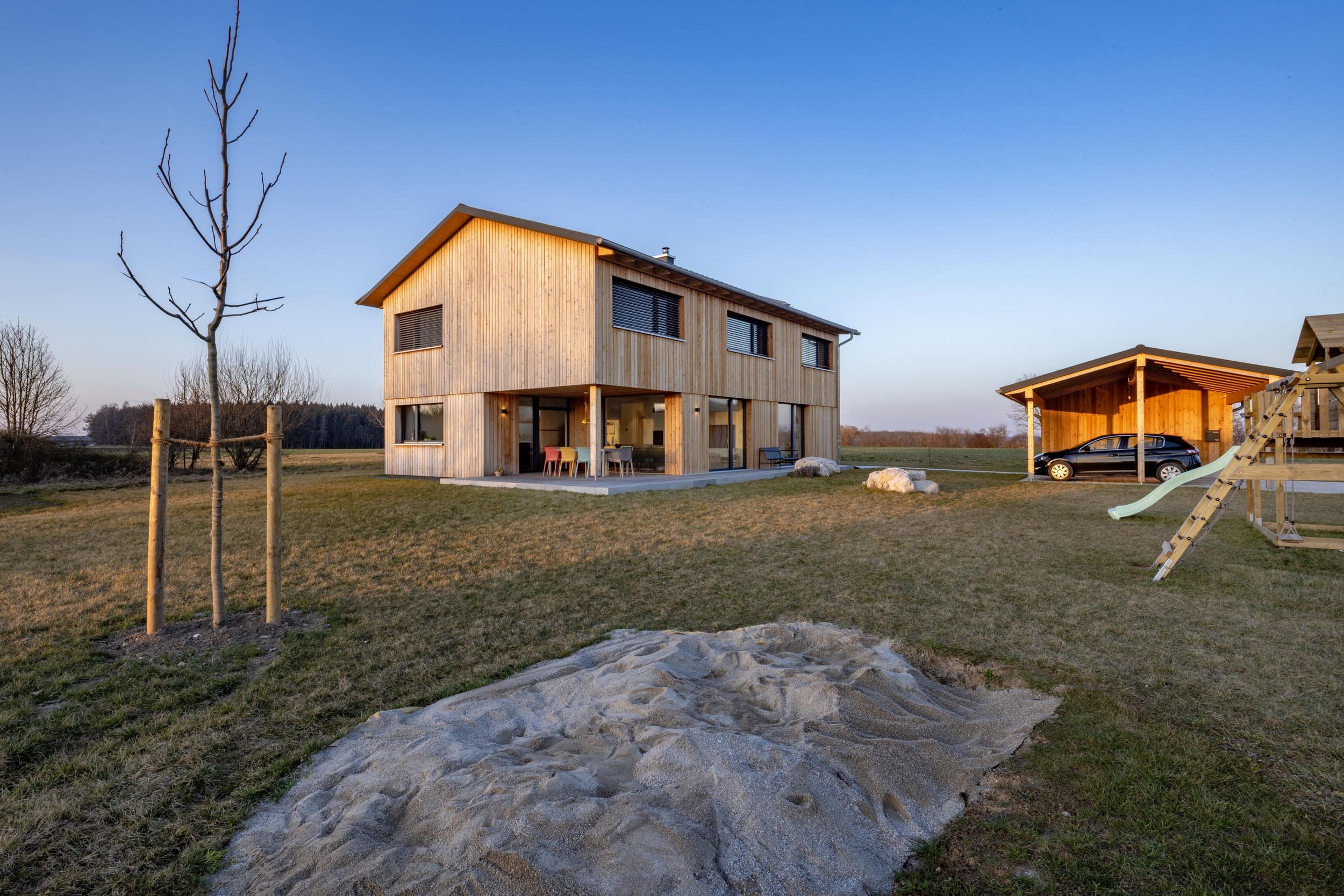 Ein modernes Holzhaus mit Terrasse steht in einem weitläufigen Garten neben einer Schaukel und einem Carport bei Sonnenuntergang.