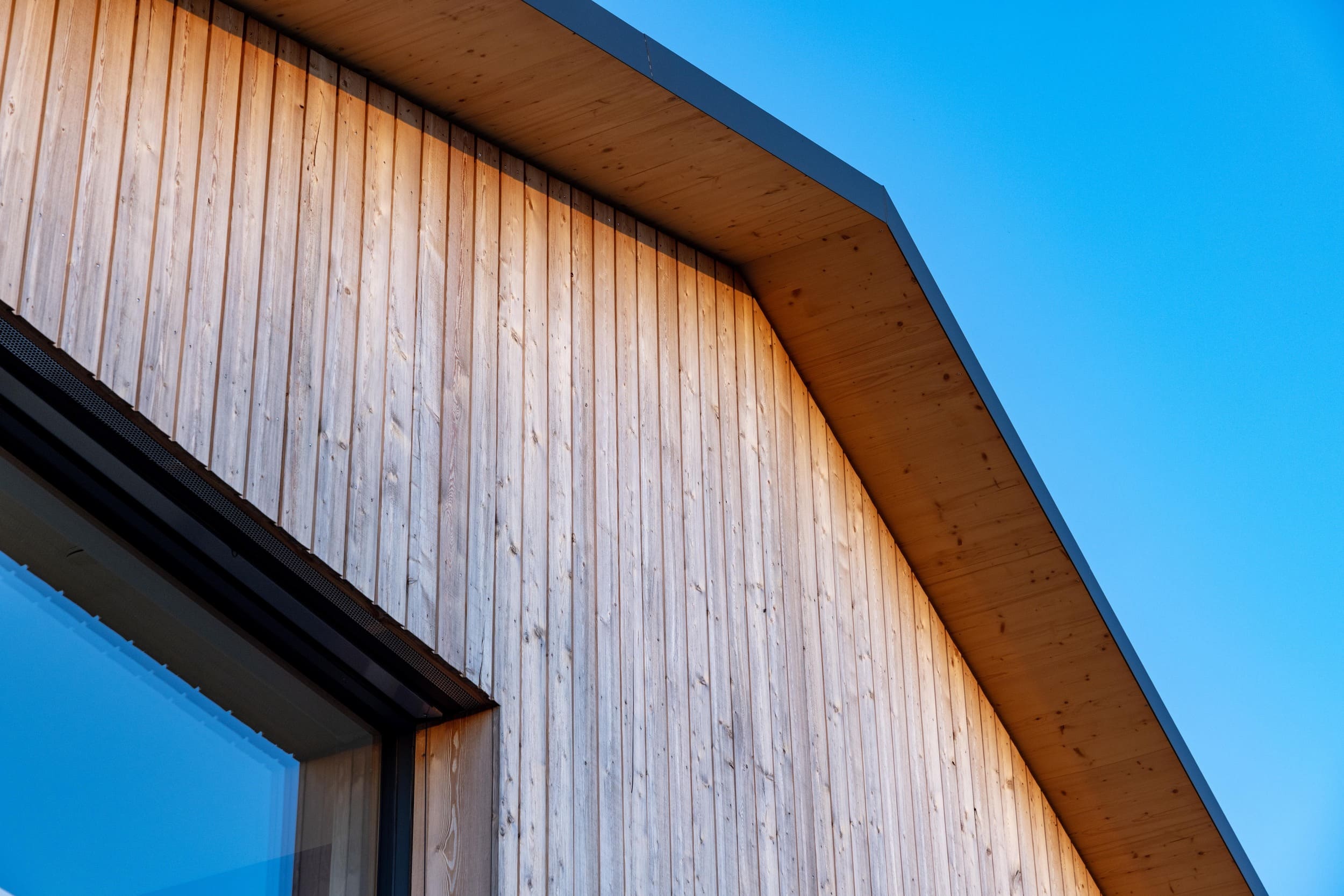 Ein modernes Holzhaus mit schrägem Dach wird von der Sonne beleuchtet, während der blaue Himmel im Hintergrund strahlt.
