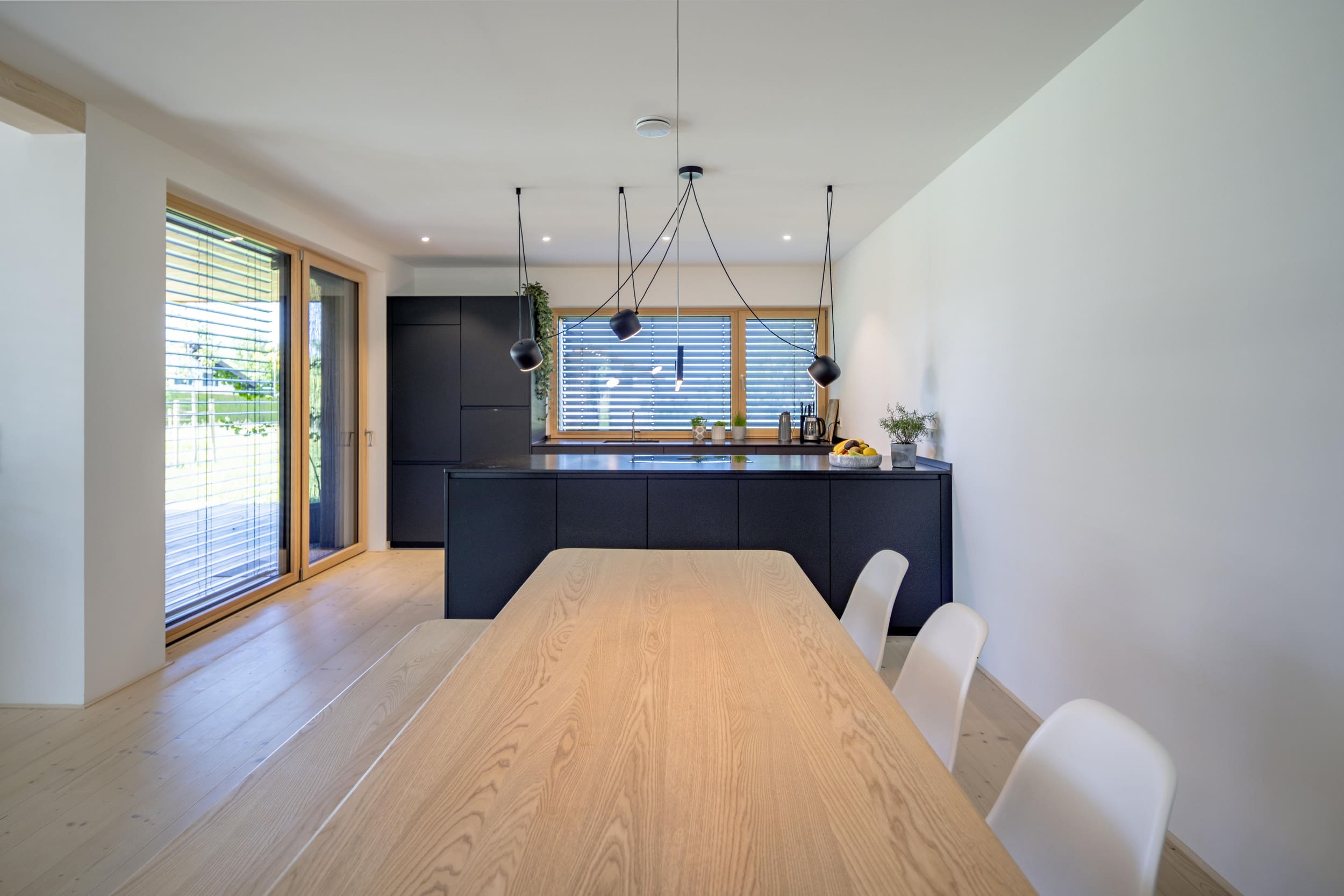 Moderner Essbereich mit Holztisch und Blick auf eine stilvolle Küche mit dunklen Schränken und eleganten Hängelampen.