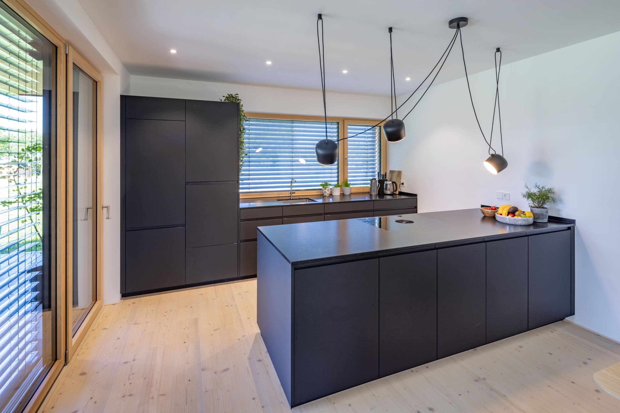 Moderne Küche mit schwarzer Insel, Hängelampen und Holzfußboden vor einer großen Fensterfront mit Jalousien.