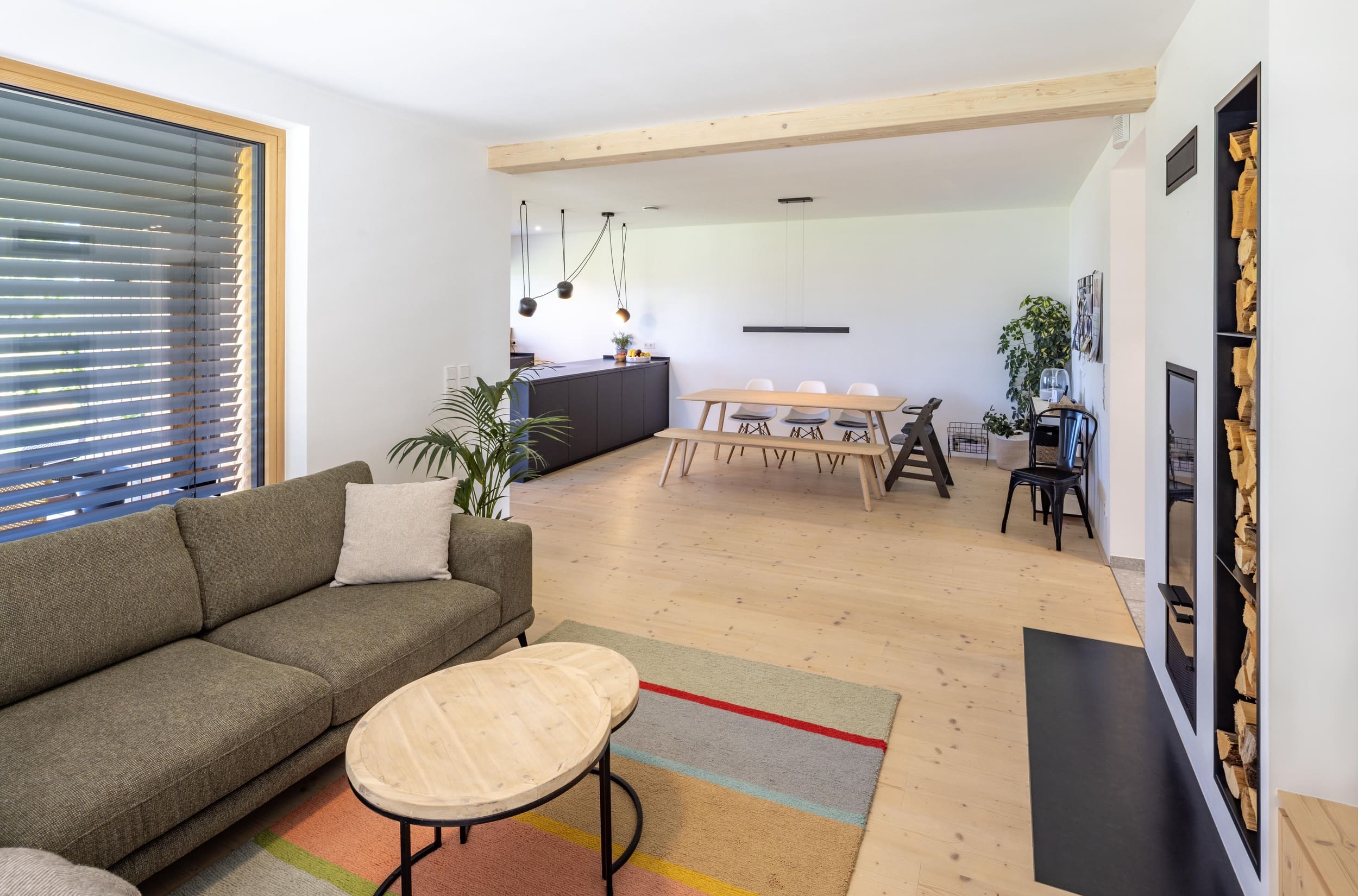 Ein modernes Wohnzimmer mit grauem Sofa, Holztisch und offener Küche, beleuchtet von Pendelleuchten, schafft eine gemütliche Atmosphäre.