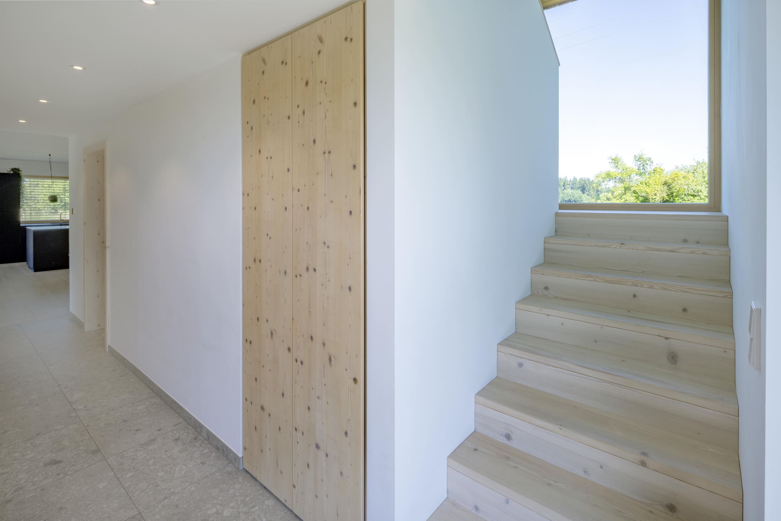 Eine helle Holztreppe führt in einem modernen Flur zu einem Fenster mit Ausblick auf grüne Bäume und blauen Himmel.