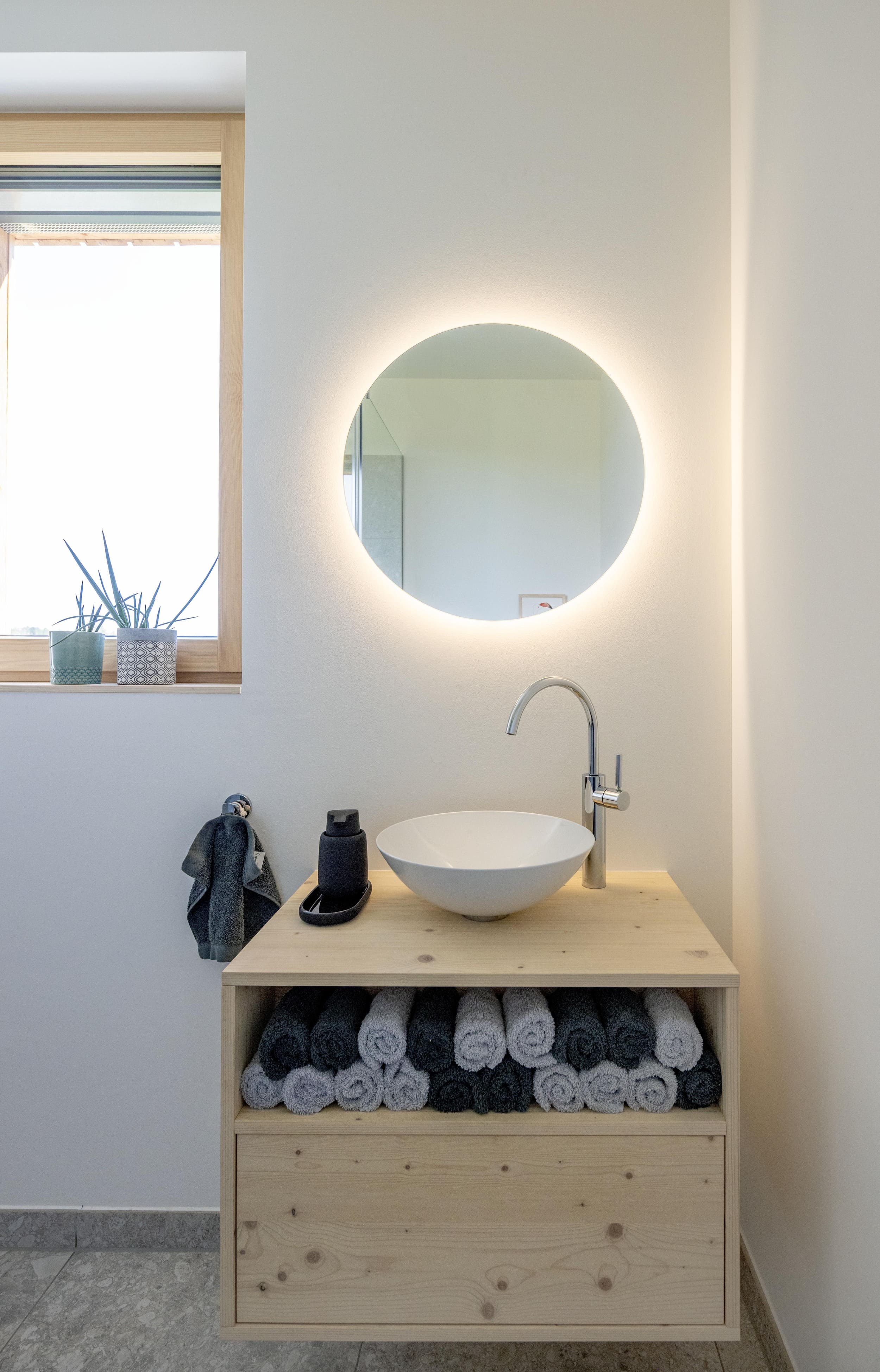 Ein minimalistisches Badezimmer zeigt einen beleuchteten runden Spiegel über einem Waschbecken mit ordentlich gestapelten Handtüchern.