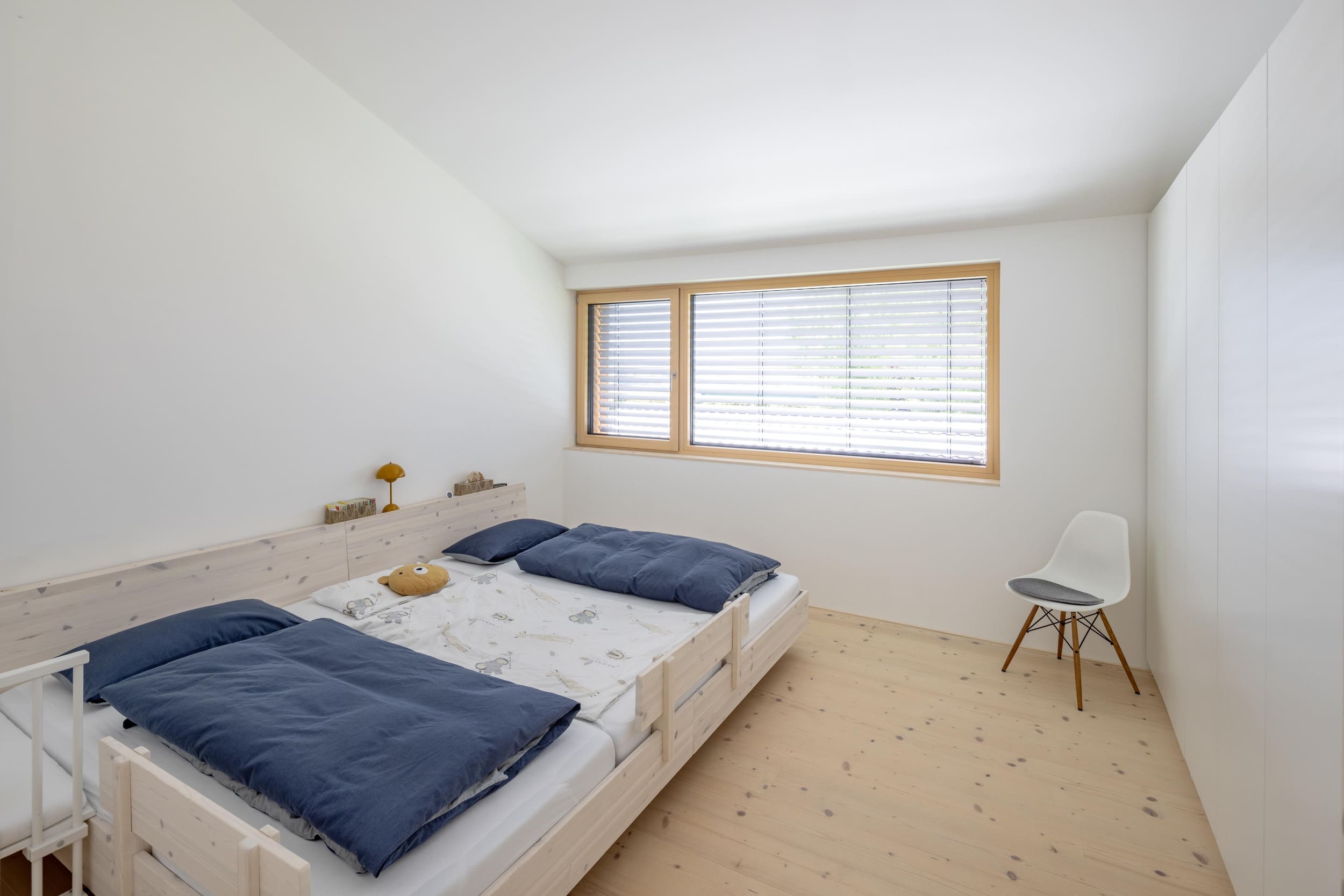 Ein schlichtes Schlafzimmer mit Holzfußboden zeigt ein Doppelbett mit blauen Decken und ein Fenster mit Jalousien.
