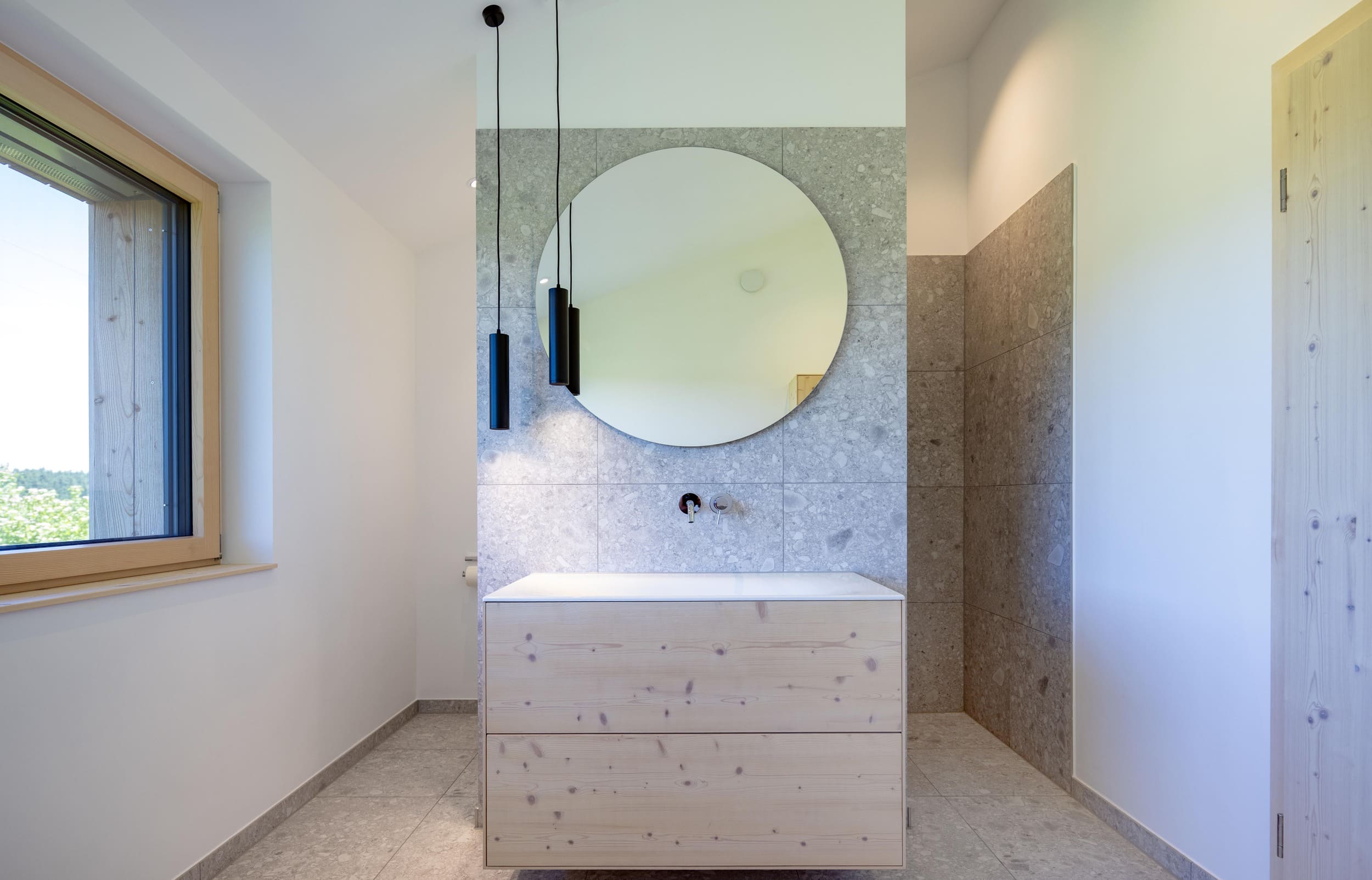 Modernes Badezimmer mit kreisförmigem Spiegel, hölzernem Waschtisch und großen Fenstern, die viel natürliches Licht hereinlassen.